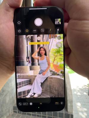 A person is holding an iPhone, which displays a photo of a young woman sitting on steps, posing for a photo. The phone's camera interface is visible, with various settings and buttons around the edges. The woman in the photo is wearing a light blue top, white pants, and white shoes with black stripes. The background is blurred, suggesting a park or outdoor setting with greenery. The hands holding the phone are visible against a soft cream background. A yellow text box with Vietnamese text is visible at the top of the screen.