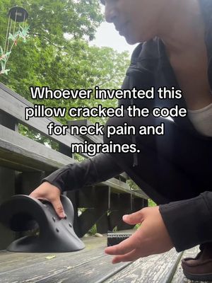 A person is holding a black neck pillow on a wooden surface. The person is partially visible, with a focus on the upper body and hands. The background includes greenery and a wooden fence. Text overlaid on the image reads: "Whoever invented this pillow cracked the code for neck pain and migraines."