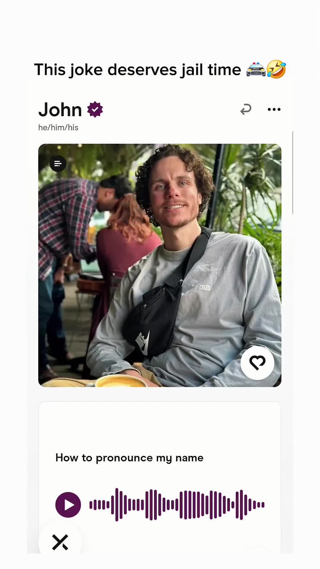 A selfie-style shot featuring a man smiling at the camera, with a background of a restaurant setting. The man is wearing a light gray long-sleeved shirt, a black fanny pack, and has curly hair. The image has overlaid text and a play button graphic with a soundwave, implying an audio element accompanies the visuals.