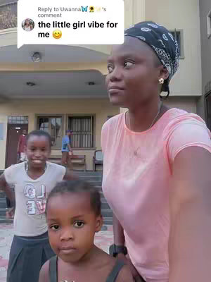 A selfie-style video cover featuring a young woman with a headscarf and two young girls in the foreground. The woman is in the right side of the image, looking toward the left. The girls are facing the camera, with one in the front. The background includes a building and a glimpse of people.