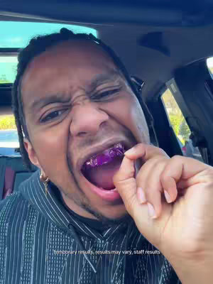 A close-up shot of a man inside a car, showing him with a look of surprise and excitement. His mouth is open, revealing purple-colored material in his teeth, with his right hand holding something near his mouth. The background suggests a daytime setting.