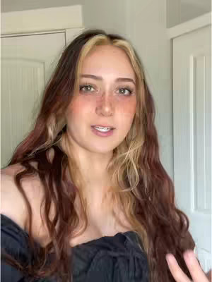 A young woman with long, wavy highlighted hair and freckles is centered in the frame. She wears a black off-the-shoulder top and appears to be speaking, with a neutral expression. The background shows partial white doors.