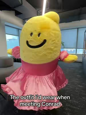 The cover image shows a person in a large, humorous costume resembling a yellow, egg-shaped character with a smiling face. The character wears a pink dress and stands in an office setting. The background includes windows overlooking city views, gray seating, and open spaces.