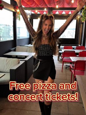 A woman with long brown hair is standing in an outdoor dining area with her arms raised in excitement. She's wearing a black top, skirt, and knee-high boots. The background includes tables, chairs, and a red awning.