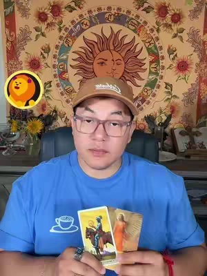 A man wearing glasses, a cap, and a blue t-shirt is holding tarot cards. The background includes a tapestry with a sun design and a floral pattern. A logo appears in the top left corner.