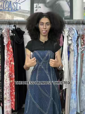 A woman with large, dark curly hair and glasses stands in front of a clothing rack. She is wearing a dark top and a blue dress, flashing a thumbs-up with both hands. Text overlayed reads "THE DRESS I ALMOST THRIFTED". The background features racks of clothes in a store setting.
