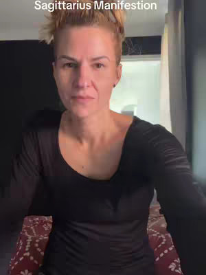 A woman with fair skin and light brown hair, pulled up in a bun, looks directly at the camera in a medium shot. The text 'Sagittarius Manifestation' is overlaid at the top, colored white. She's wearing a black long-sleeved shirt against a dark background. The lighting appears to be natural, as if in a room with light coming in from the left.