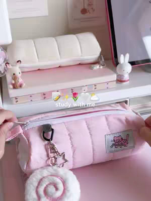 A close-up shot featuring a pink pencil case, a decorative shelf with items like a white bunny figurine and a stack of notebooks. The color palette is dominated by pastels, with the pencil case in focus, held open, and a white, swirly charm attached.