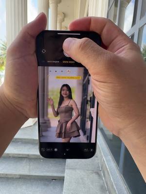 A person's hands holding an iPhone, displaying a portrait photo of a woman. The phone's screen shows the camera interface, with the shutter button highlighted. The woman in the photo is smiling and waving. The background shows a building's entrance.