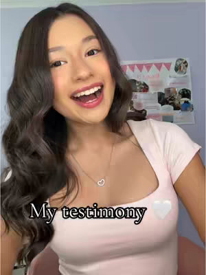 A young woman with long, wavy dark hair smiles widely, looking directly at the viewer. She wears a light pink square-neckline top and a delicate necklace with a heart pendant. The background is a blurred mix of colors, with a blurred image visible behind her. Text overlayed on the image says 'My testimony' in a casual font, with a small white heart symbol next to it.