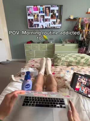 The cover image shows a first-person perspective of a person lying in bed with their legs in the foreground. The person is wearing white socks and their legs are slightly bent. In front of them is a laptop with a colorful sticker on the cover. A bottle of lotion and a box of tissues are to the left. On the wall behind the feet is a television displaying a grid of colorful images of women. The overall composition is casual and intimate, suggesting a relaxed, morning routine.