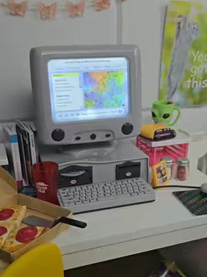 A miniature recreation of a 1990s computer setup with a pizza, books, and an alien-themed mug. The setup features an old-style monitor displaying a map and a keyboard on a white desk. The lighting is bright, and the scene is set up with attention to detail, evoking nostalgia.