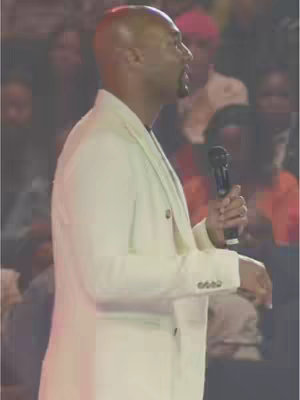A man in a white suit speaks into a microphone, standing on a stage with an audience blurred in the background.