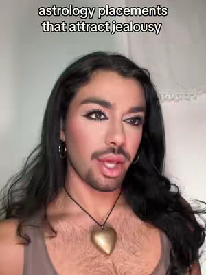 A person with long dark hair, full makeup, and a beard is centered in the frame. The background seems to be a plain white wall. The person is wearing a dark necklace with a gold heart-shaped pendant and a tan top. Text in white across the top of the image reads: "astrology placements that attract jealousy"