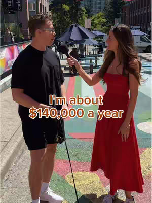 A street interview scene. A man in black shorts and t-shirt and glasses is being interviewed by a woman in a red dress. The woman is holding a microphone. The background includes buildings, trees, and painted pavement.