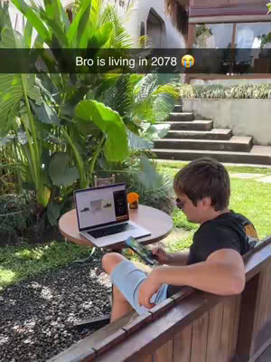 A young man sits outside at a round table with a laptop open, likely using a web scraping tool. He is holding another electronic device. Lush greenery and a building with stairs in the background suggest a pleasant outdoor setting.