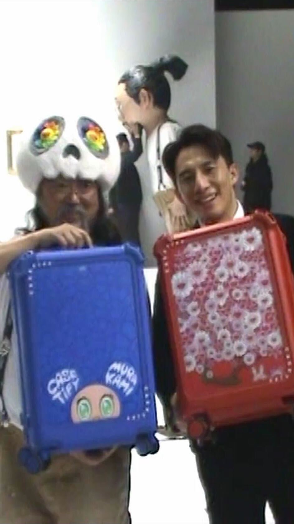 An image captured at an art exhibition, featuring two men holding CASETiFY suitcases. The background is a bright white wall. The men are smiling and facing forward. One man is wearing a panda-skull hat and holding a blue suitcase; the other is wearing a blazer and holding a red suitcase. Artwork and figures are visible in the blurry background. The overall composition is well-lit.