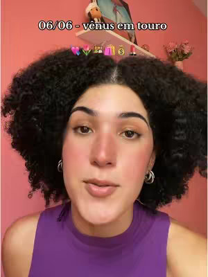A woman with curly dark hair and a purple top looks directly at the camera. Above her head, a text overlay states '06/06 - vênus em touro,' with associated emojis. The background is a light pink wall.