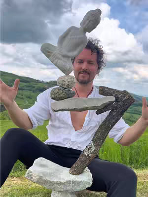 The cover image showcases a man sitting outdoors with an arrangement of stacked objects in front of him. He smiles towards the viewer with arms open in a gesture that mirrors the balance of the objects. The background is filled with lush green hills and a sky dotted with clouds.