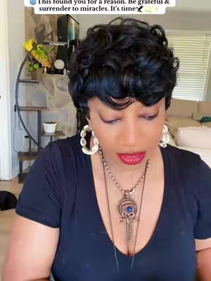 A woman with dark, curly hair looks down, wearing large hoop earrings and a necklace; the background is a living room.