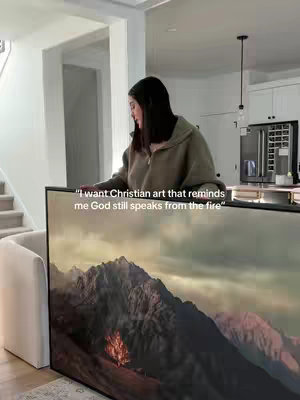 A woman stands behind a large framed artwork in a home setting, displaying artwork that depicts a mountain scene with a fire in the foreground. Text overlays are placed near the top of the frame.