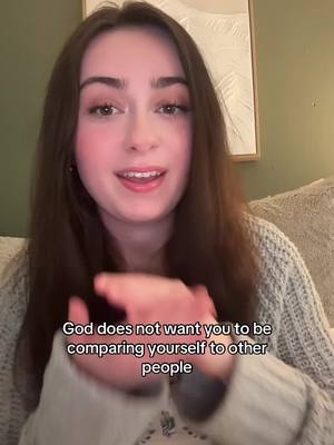 A young woman with long brown hair is centered in the frame, smiling and looking directly at the camera. She wears a light gray sweater. The background includes a white framed artwork and a soft, neutral-toned setting. Text overlayed at the bottom states, "God does not want you to be comparing yourself to other people."