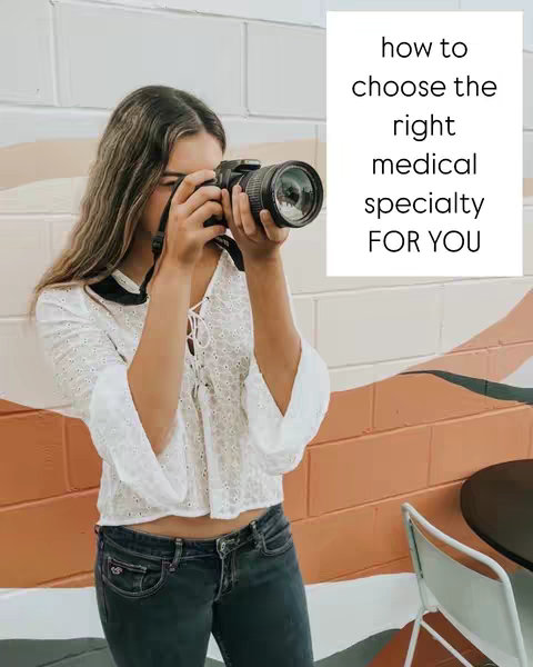 A young woman, wearing a white blouse and dark jeans, is holding a camera up to her face as if taking a photo. She is standing in front of a wall with a white section on the right, which has text on it. The wall to the left of the text is a mix of colors and shapes including white, brown, and orange.