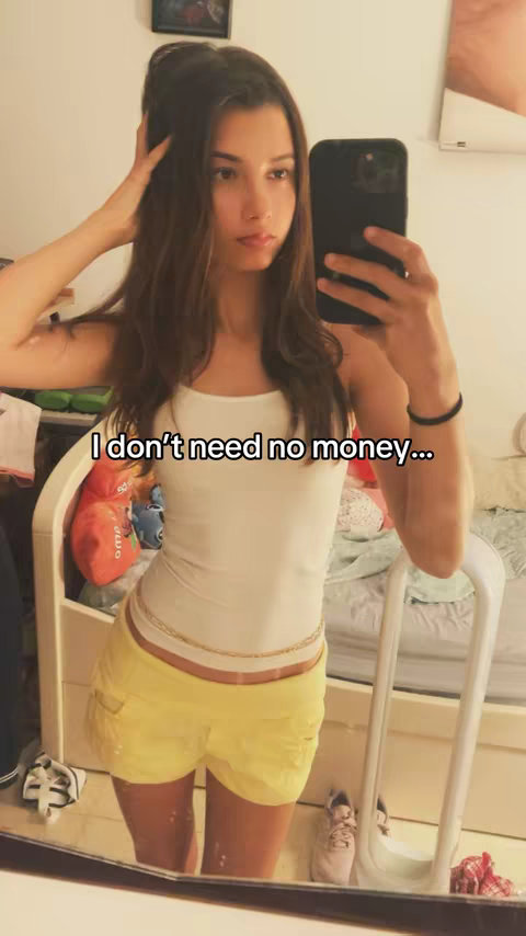 A young woman is taking a mirror selfie in a bedroom setting. She is wearing a white tank top, yellow shorts, and a gold chain belt. She is holding a black phone in front of her face, partially obstructing her view. The text "I don't need no money..." is overlaid on the image in white font. The mirror captures the reflection of the room, including clothing and a partly visible window.