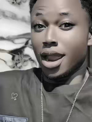 A close-up shot features a young, light-skinned Black man with short, dark hair. His mouth is open in an expression of surprise or excitement, showing his teeth slightly. The background is a blurred mix of neutral tones. He is wearing a dark gray t-shirt and a silver chain is visible around his neck. The lighting appears to be soft and diffused, enhancing the slightly blurred aesthetic.
