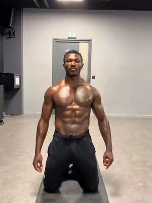 A muscular man with sweat glistening on his skin kneels on an exercise mat. He's shirtless, wearing only black shorts, and is positioned in a fitness environment with a workout mat on the floor.