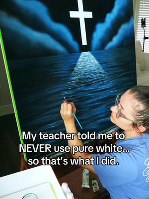 An oil painting in progress, depicting a cross in a night sky illuminating the sea with a beam of light. A woman is painting the artwork, wearing glasses and a blue shirt. The green side of the art is visible.