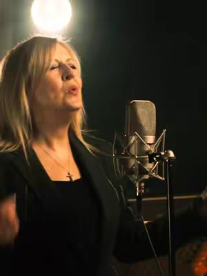 A woman with blonde hair is singing into a microphone, lit by a bright overhead light. She is wearing a dark outfit and a cross necklace. The background is dark, and a microphone stand is visible. The overall composition focuses on the performer during a musical performance.