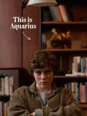 A person with short, curly brown hair looks directly at the viewer with a neutral expression. They are wearing a brown corduroy jacket over a gray sweater. The background includes a bookshelf filled with books and a warm-toned wall, with text overlay reading 'This is Aquarius' beside text and an arrow pointing to the person.