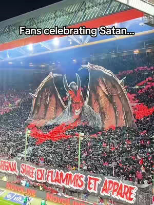 The cover image shows a stadium filled with people, many holding up red and black banners. In the center, a giant image of a winged, devil-like figure with horns is displayed. The image is well-lit, with focus on the crowd, the giant banner and the message written in large letters. The overall color scheme is dark, with bright red accents.