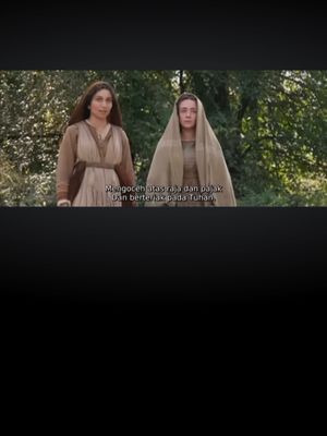 Two women stand in a natural setting, framed from the waist up. The woman on the left wears a brown dress, and the woman on the right wears a white head covering and a dress. Between them is a text overlay.