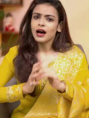 A woman with long dark hair wearing a yellow and white patterned outfit is looking towards the camera with her mouth open, suggesting she is speaking. She has a bindi on her forehead.