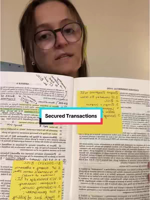 A woman wearing glasses is the focus, holding open a law book with handwritten notes and Post-it notes. The image has a warm color tone and is lit from from above.