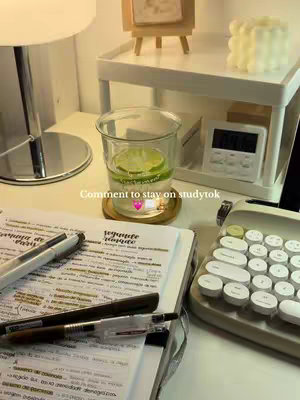 The image shows a well-lit desk setup, likely for studying, with a notebook open to notes highlighted in yellow, pens, a glass with lemon in it, and a retro style dial-pad looking keyboard. A timer, small picture frame and candle can be seen in the background.