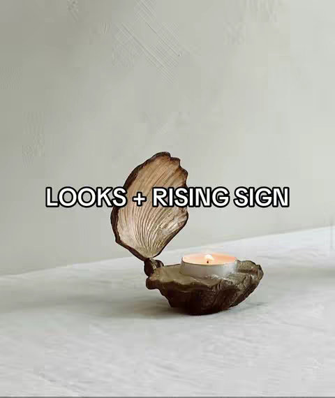 A still life setup features a decorative seashell shaped candle holder on a white surface against a light gray background. The candle holder in the center resembles a partially open clam, composed of a rough, earth-toned material. Inside the holder, a lit tealight candle provides warm light. The text 'LOOKS + RISING SIGN' is overlaid on the image.