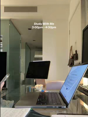An interior shot showing a brightly lit workspace. A laptop is open on a glass desk with a partially visible monitor to the right and an iPad or tablet propped up on a stand behind it. The background includes an open doorway, interior hallway. The text overlay indicates a study session.