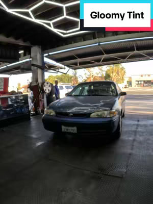 The image shows a dark blue car in an automotive shop with a window tinting service being performed. The car is positioned in the center of the frame, with the front facing the viewer. Above is a white and red text overlay reading "Gloomy Tint."