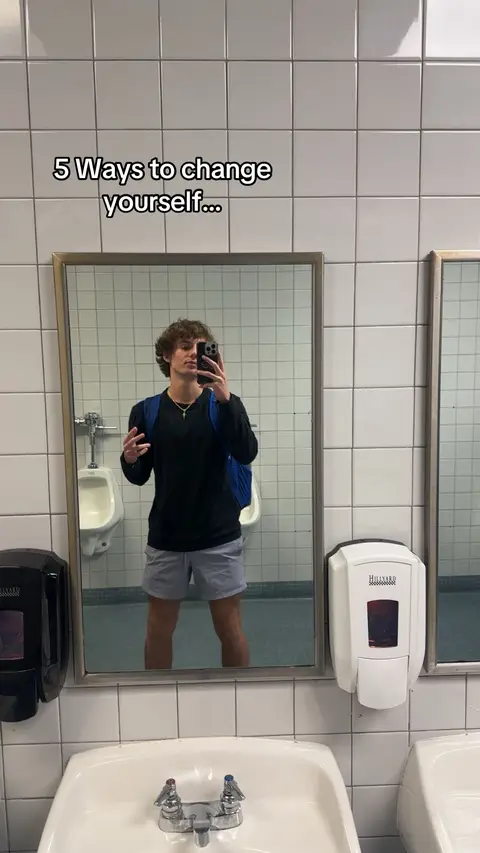 A young man takes a mirror selfie in a public restroom. The background is tiled with white square tiles. The man is wearing a black long-sleeve shirt and grey shorts, carrying a blue backpack. The text '5 Ways to change yourself...' is overlaid at the top.