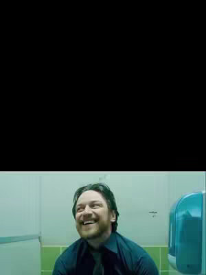 A man smiles broadly while looking upwards in a public restroom. The background consists of green tiles and a blue paper towel dispenser.