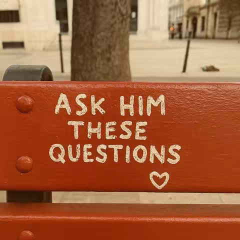 A close-up shot of a wooden bench painted red. The words "ASK HIM THESE QUESTIONS" are stenciled on the bench in stylized white letters. A small white heart graphic is painted below the text. The background is blurred, suggesting an outdoor setting.