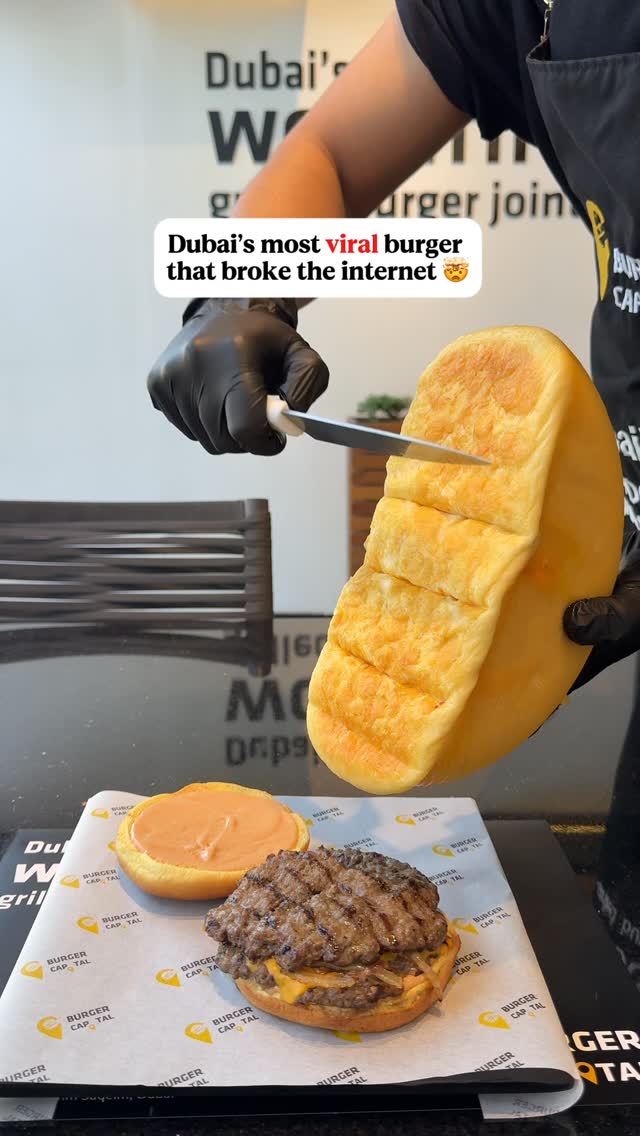A top-down shot presents a freshly prepared burger, meticulously arranged on a branded paper. A large, golden-brown loaf of bread, seemingly cheese, is being sliced by a person wearing black gloves. The top of the burger is cut open, revealing the patty and the cheese