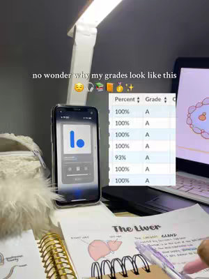 A well-lit, close-up shot of a study desk. A smartphone displaying an app is propped up in front. A laptop and notes on anatomy are visible on the desk. The image is well composed and the colors are natural.