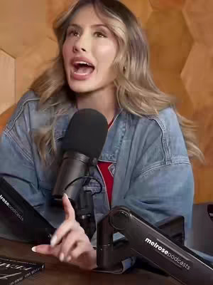 A woman with long, wavy blonde hair speaks passionately into a microphone. She's wearing a denim jacket over a red top. Two black microphone stands featuring "melrose podcasts" are in front of her. The background shows a tan, geometric wooden wall.
