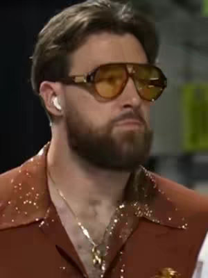 A man wearing sunglasses and a sparkly collared shirt is shown from the chest up. He has a beard and an earpiece. The background is blurred.