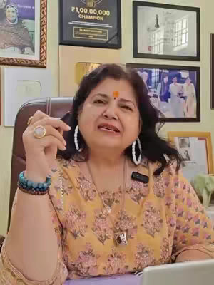 A woman with dark hair and a bindi sits in a chair, looking towards the camera and talking. She has a floral top and large hoop earrings. The background features framed photos and certificates on the wall, suggesting a professional setting.