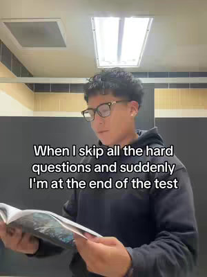 A young man with black hair and glasses is standing in a bathroom, holding a book and looking down at it. The text overlayed on the image reads "When I skip all the hard questions and suddenly I'm at the end of the test."